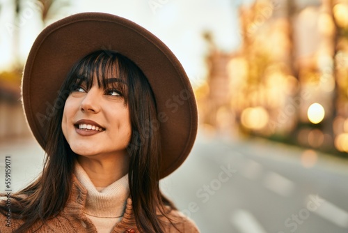 Brunette woman wearing winter hat smiling outdoors at the city on sunset