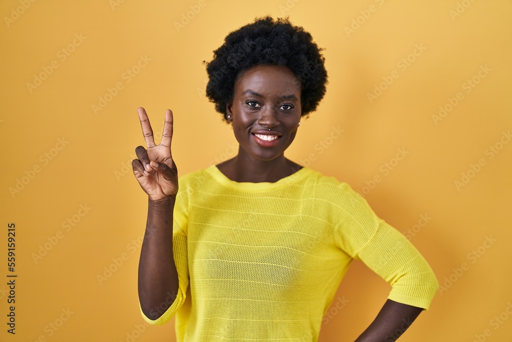 African young woman standing over yellow studio smiling looking to the camera showing fingers doing victory sign. number two. African young woman standing over yellow studio smiling looking to the camera showing fingers doing victory sign. number two.