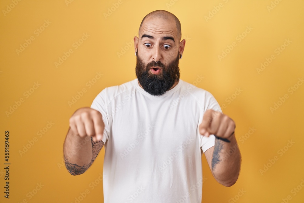 Young hispanic man with beard and tattoos standing over yellow background pointing down with fingers showing advertisement, surprised face and open mouth Young hispanic man with beard and tattoos standing over yellow background pointing down with fingers showing advertisement, surprised face and open mouth