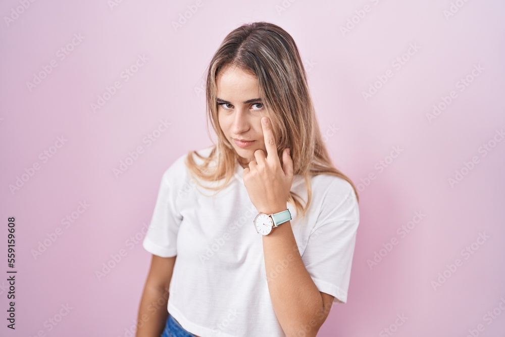 Young blonde woman standing over pink background pointing to the eye watching you gesture, suspicious expression Young blonde woman standing over pink background pointing to the eye watching you gesture, suspicious expression