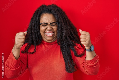 Plus size hispanic woman standing over red background excited for success with arms raised and eyes closed celebrating victory smiling. winner concept. © Krakenimages.com