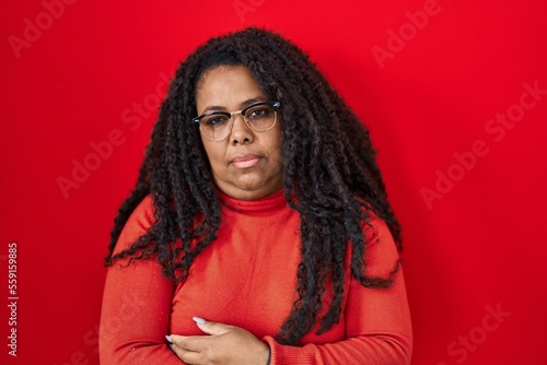 Plus size hispanic woman standing over red background looking sleepy and tired, exhausted for fatigue and hangover, lazy eyes in the morning.