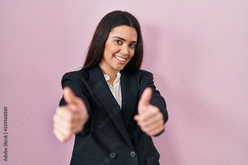 Young brunette woman wearing business style over pink background approving doing positive gesture with hand, thumbs up smiling and happy for success. winner gesture. Young brunette woman wearing business style over pink background approving doing positive gesture with hand, thumbs up smiling and happy for success. winner gesture.