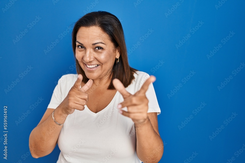 Fototapeta premium Hispanic mature woman standing over blue background pointing fingers to camera with happy and funny face. good energy and vibes.