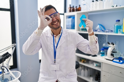 Young hispanic man with beard working at scientist laboratory holding blue ribbon smiling happy doing ok sign with hand on eye looking through fingers
