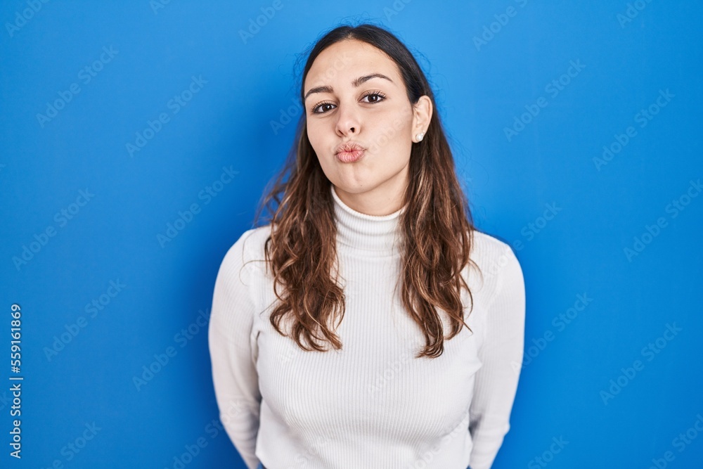 Obraz premium Young hispanic woman standing over blue background looking at the camera blowing a kiss on air being lovely and sexy. love expression.