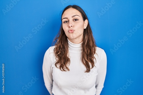 Young hispanic woman standing over blue background looking at the camera blowing a kiss on air being lovely and sexy. love expression.