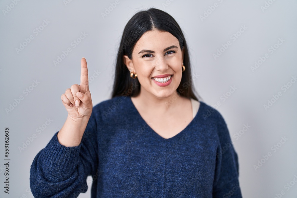 Young brunette woman standing over isolated background showing and pointing up with finger number one while smiling confident and happy. Young brunette woman standing over isolated background showing and pointing up with finger number one while smiling confident and happy.