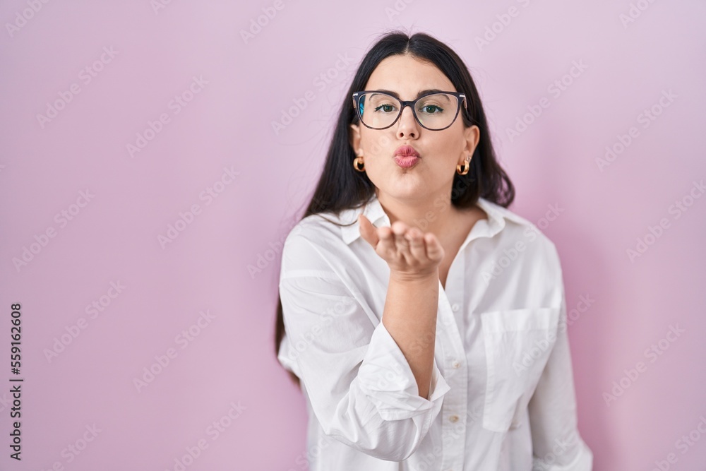 Young brunette woman standing over pink background looking at the camera blowing a kiss with hand on air being lovely and sexy. love expression. Young brunette woman standing over pink background looking at the camera blowing a kiss with hand on air being lovely and sexy. love expression.
