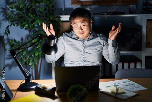 Young chinese man working using computer laptop at night clueless and confused expression with arms and hands raised. doubt concept.