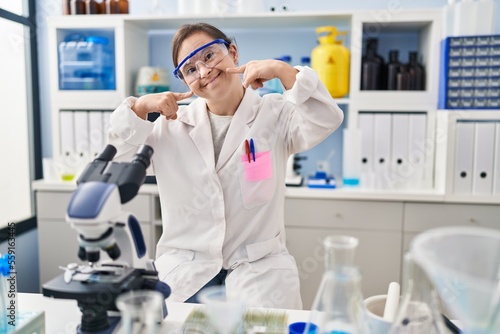 Hispanic girl with down syndrome working at scientist laboratory smiling cheerful showing and pointing with fingers teeth and mouth. dental health concept.