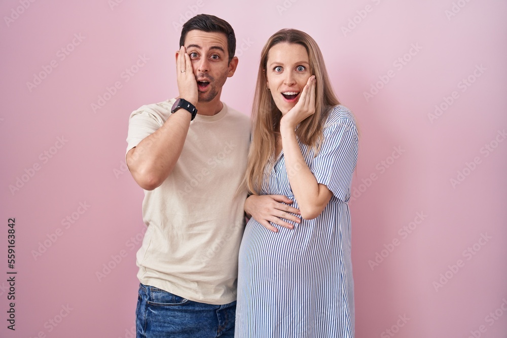 Naklejka premium Young couple expecting a baby standing over pink background afraid and shocked, surprise and amazed expression with hands on face