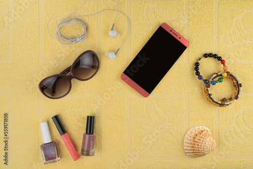Women's accessories for a beach holiday. A smartphone, jewelry and cosmetics, sun glasses, headphones and a shell in the background of a yellow terry towel.