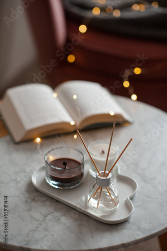 Cozy home atmosphere with liquid fragrance in glass bottle and sticks with open paper book over glow lights on marble table in living room close up. Winter holiday season.