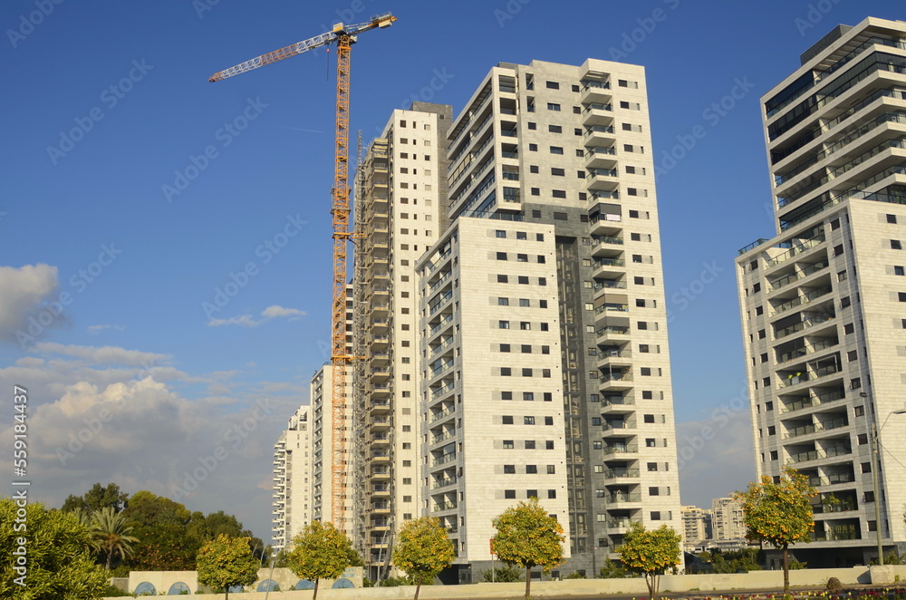 Construction of a residential building. Construction crane, tree with orange fruits. Garden next to an apartment building. Concept: buying a home, investment, mortgage, loan, price increase, real esta Construction of a residential building. Construction crane, tree with orange fruits. Garden next to an apartment building. Concept: buying a home, investment, mortgage, loan, price increase, real esta