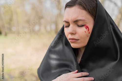 Young caucasian woman in black headscarf with closed eyes and bloody tears. Crying grieving widow in desperation.