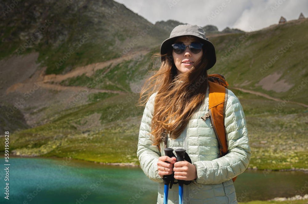 Naklejka premium Portrait of a smiling woman resting during a mountain hike. A woman with a backpack enjoys the view, standing at the lake and at the foot of the mountain