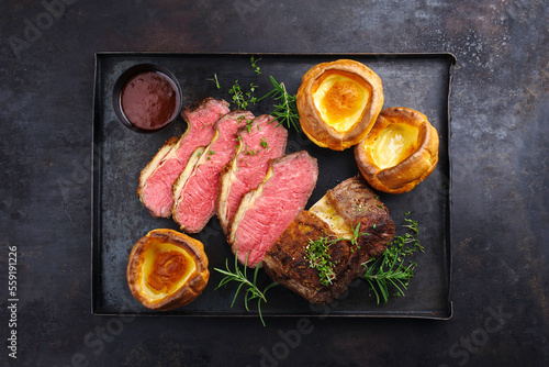 Traditional Commonwealth Sunday roast beef sliced with Yorkshire pudding and red wine sauce served as top view on a rustic black metal tray