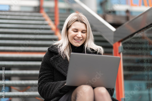 una donna felice lavora a distanza in una strada cittadina, usa il laptop e si siede sulle scale. Libero professionista femminile che lavora online al giorno d'estate. Lavoro a distanza e freelance photo