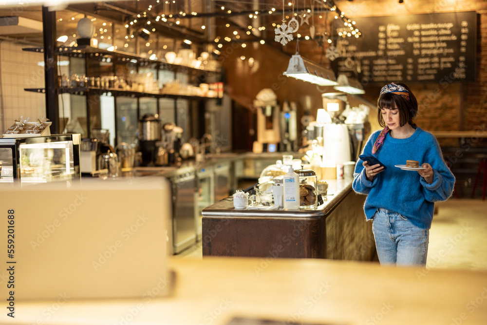 Young stylish woman using phone while carrying her order at modern coffee shop. Concept of modern lifestyle and digital work at cafe