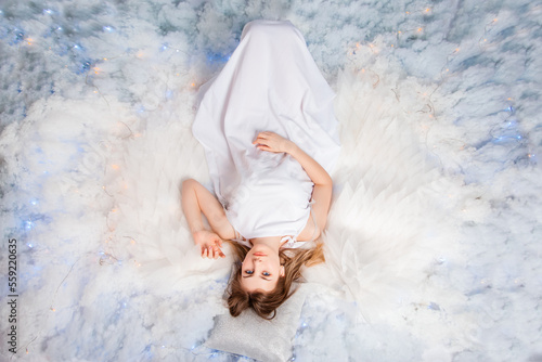 Top view, an attractive girl in a white tunic with large white wings behind her back poses while lying in white glowing clouds. photo