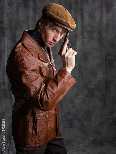 young man, a retro secret agent, a guy in a brown leather jacket and a flat cap with a gun in his hand. kgb agent. Posing in the studio on a gray background