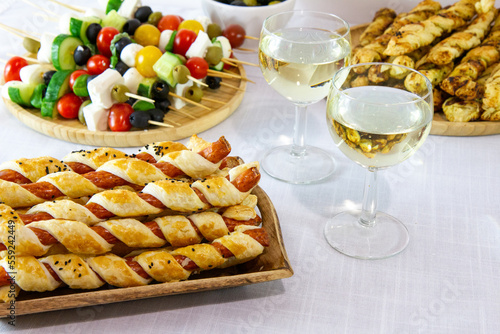 Snacks for New Year's Eve party or carnival: sausages in puff pastry, puff pastry sticks and pesto, skewers with tomatoes, olives and cheese. Next to the glasses with white wine