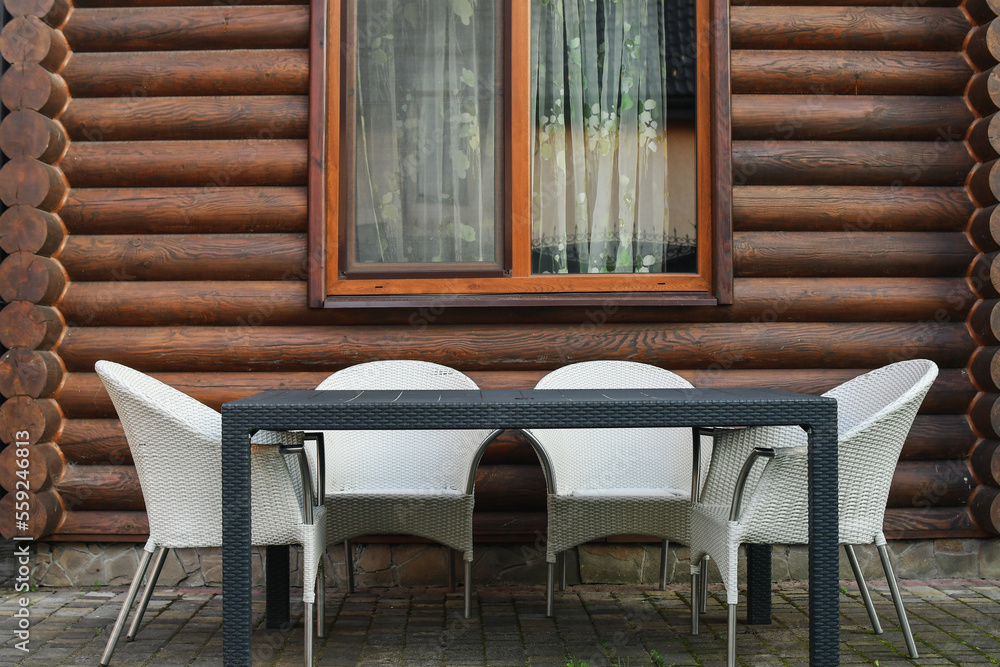 Three light rattan chairs and a table next to the wooden log house outdoors.