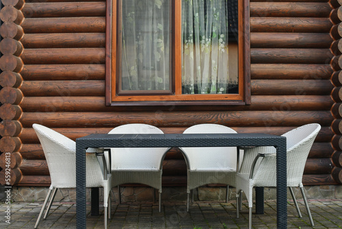 Three light rattan chairs and a table next to the wooden log house outdoors.