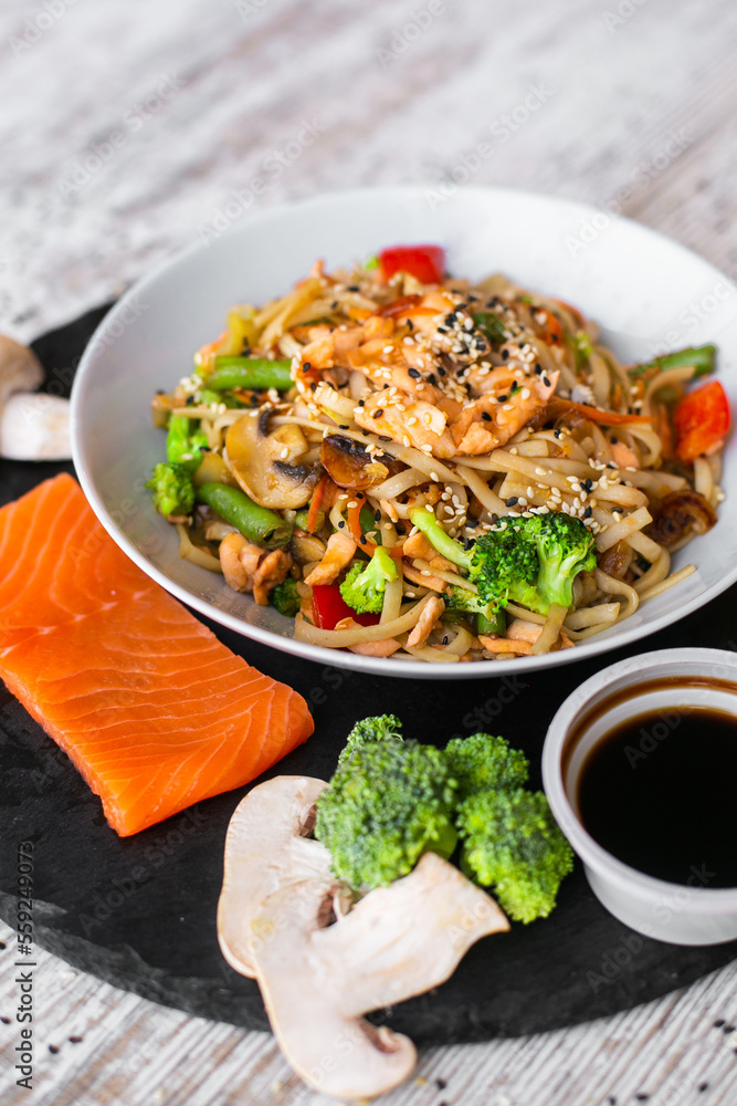 Wok, noodles, udon with salmon, on black plate and a wooden white background top view