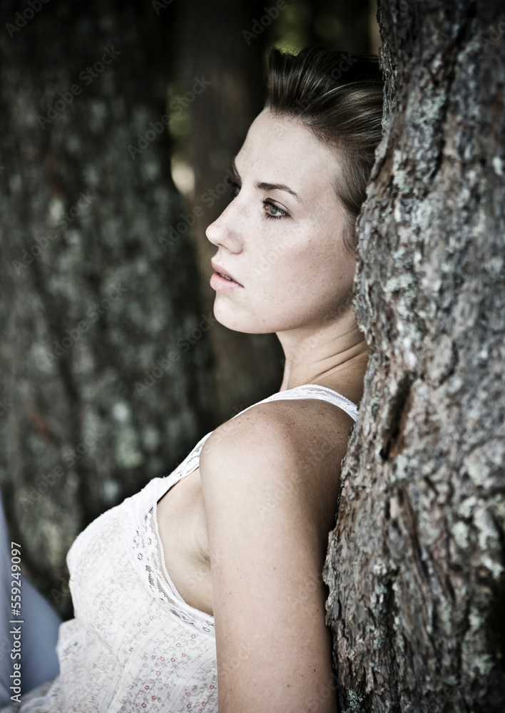 A young woman in a white tank top rests with her back against a tree. A young woman in a white tank top rests with her back against a tree.
