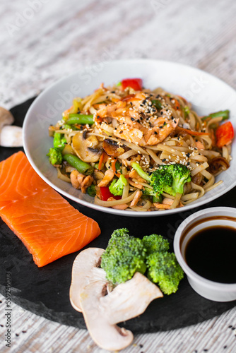 Wok, noodles, udon with salmon, on black plate and a wooden white background top view