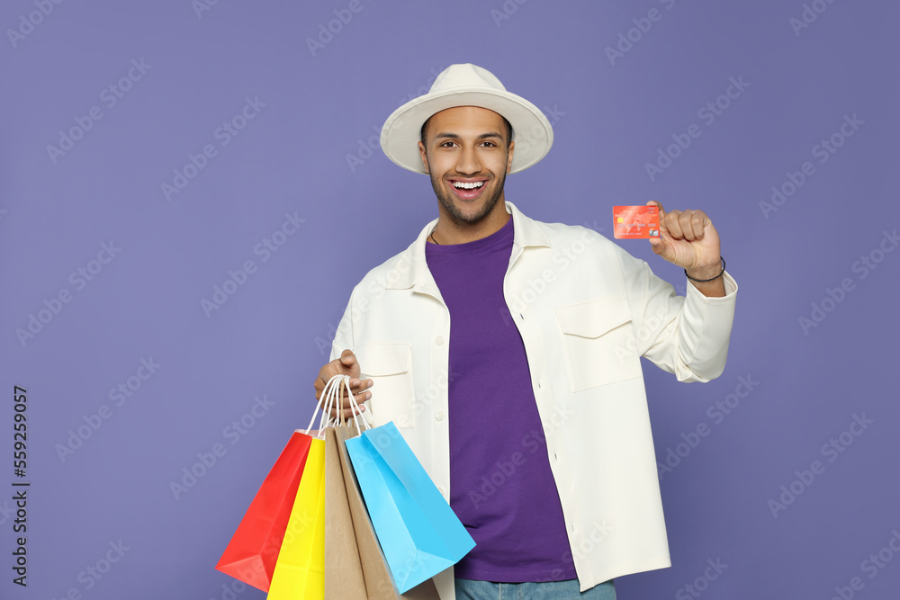 Happy African American man in hat with shopping bags and credit card on purple background Happy African American man in hat with shopping bags and credit card on purple background