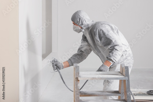 Decorator in protective overalls painting wall with spray gun indoors © New Africa