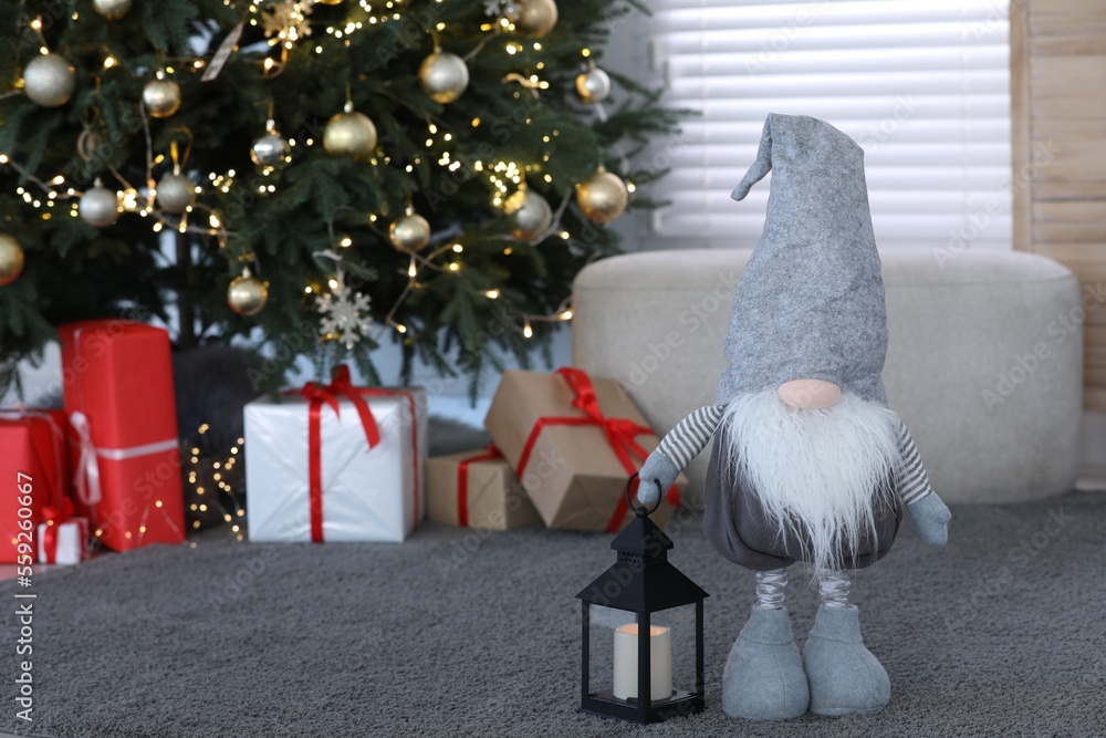 Obraz premium Cute Scandinavian gnome with lantern candle holder near Christmas tree on carpet in room. Space for text