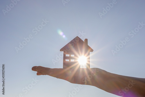 Photography Hand holding wooden house toy against blue sunny sky with sun rays