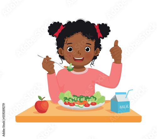 Cute little African girl eating broccoli healthy vegetables with fork showing thumb up gesture
