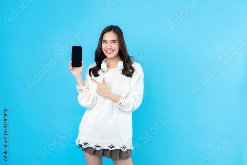 Beautiful young white Asian woman with long hair, casual wear, wearing white wireless headphones. In her hand she is pointing at a smartphone with a black screen. on the blue background in the studio.