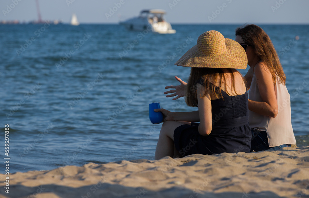 Two young women are resting on the beach - two friends are sitting on the beach, drinking drinks, talking to each other. Vacation at sea.