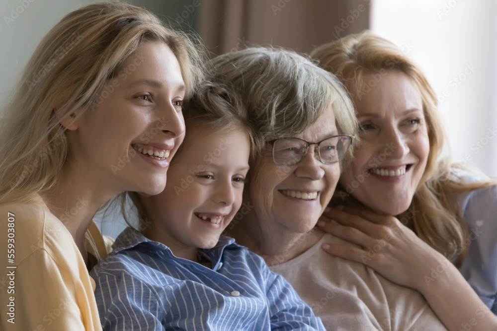 Happy pretty girls and mature senior women of four generations sitting close together, posing, looking away with toothy smiles, laughing, hugging, enjoying family leisure time Happy pretty girls and mature senior women of four generations sitting close together, posing, looking away with toothy smiles, laughing, hugging, enjoying family leisure time