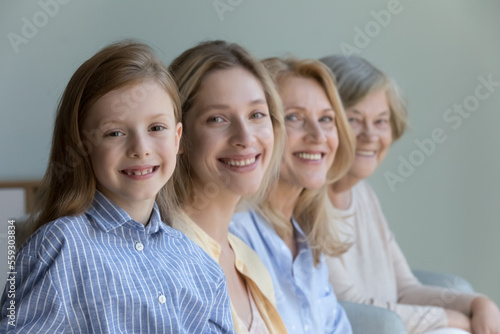 Happy beautiful cute little kid girl looking at camera, smiling, posing with young mother, mature cheerful grandma, positive great grandmother standing behind. Female generation, family portrait