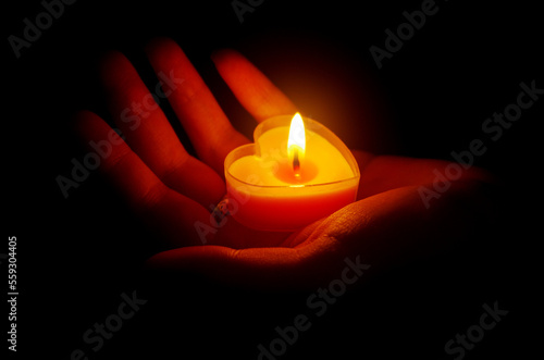 A hand holding a burning candle in the shape of a heart. Dark background.