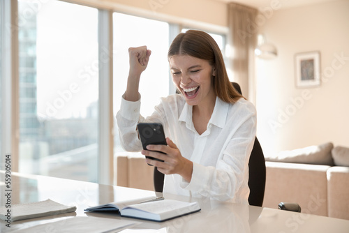 Fototapete Lucky woman sits at desk holds phone get special offer by sms feels happy, receive huge discount, fantastic news, scream with joy, enjoy moment of auction on-line lottery betting win