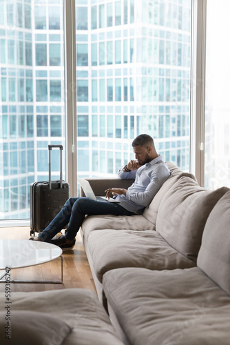 Millennial serious pensive African businessman sit on sofa working on laptop stay in luxury hotel on business trip, skyscraper buildings city view through panoramic window, vertical shot. Lifestyle
