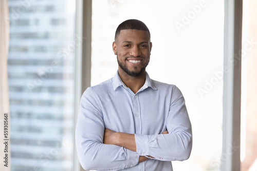 Millennial successful businessman standing in modern skyscraper office smile staring at camera looks confident. Portrait of business owner, company boss pose with arms crossed on chest at workplace
