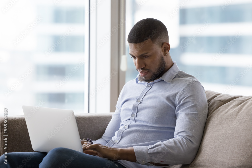 Focused African man working or studying using computer sits on sofa at modern home. Millennial American guy spend leisure on internet, typing text on laptop keyboard, buy, make order or do telework Focused African man working or studying using computer sits on sofa at modern home. Millennial American guy spend leisure on internet, typing text on laptop keyboard, buy, make order or do telework