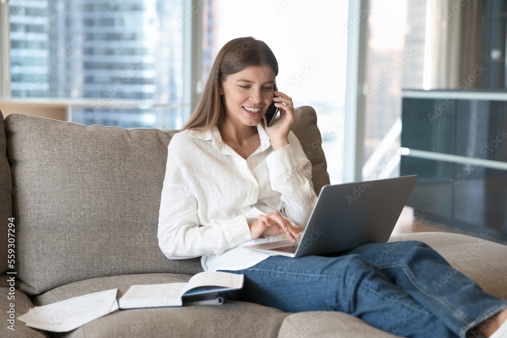Attractive young woman rests on sofa at home use laptop, holds smartphone enjoy remote conversation to friend on speaker phone. Young generation and modern tech usage. Call, communication, helpline Attractive young woman rests on sofa at home use laptop, holds smartphone enjoy remote conversation to friend on speaker phone. Young generation and modern tech usage. Call, communication, helpline