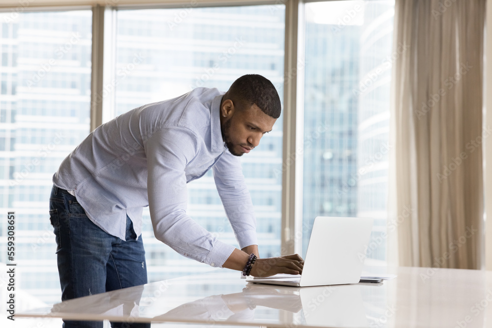 Young African businessman texting e-mail to client standing leaned over workplace desk, typing answers use laptop looks focused, finish work in modern skyscraper office. Tech, new software, lifestyle Young African businessman texting e-mail to client standing leaned over workplace desk, typing answers use laptop looks focused, finish work in modern skyscraper office. Tech, new software, lifestyle