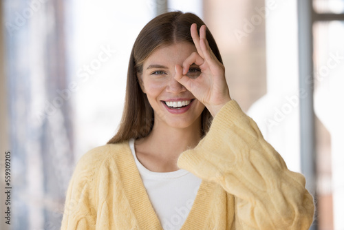 Happy young woman makes circle gesture with finger smile staring at camera, head shot. Eyesight and vision care, satisfied client of medical check up, eye-care services, myopia prevention treatment