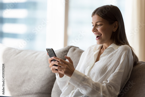 Surprised happy woman sit on sofa at home, looking at smart phone screen read good sms news feels amazed, winning in online lottery, betting, gambling, celebrate success, get sale and discounts notice